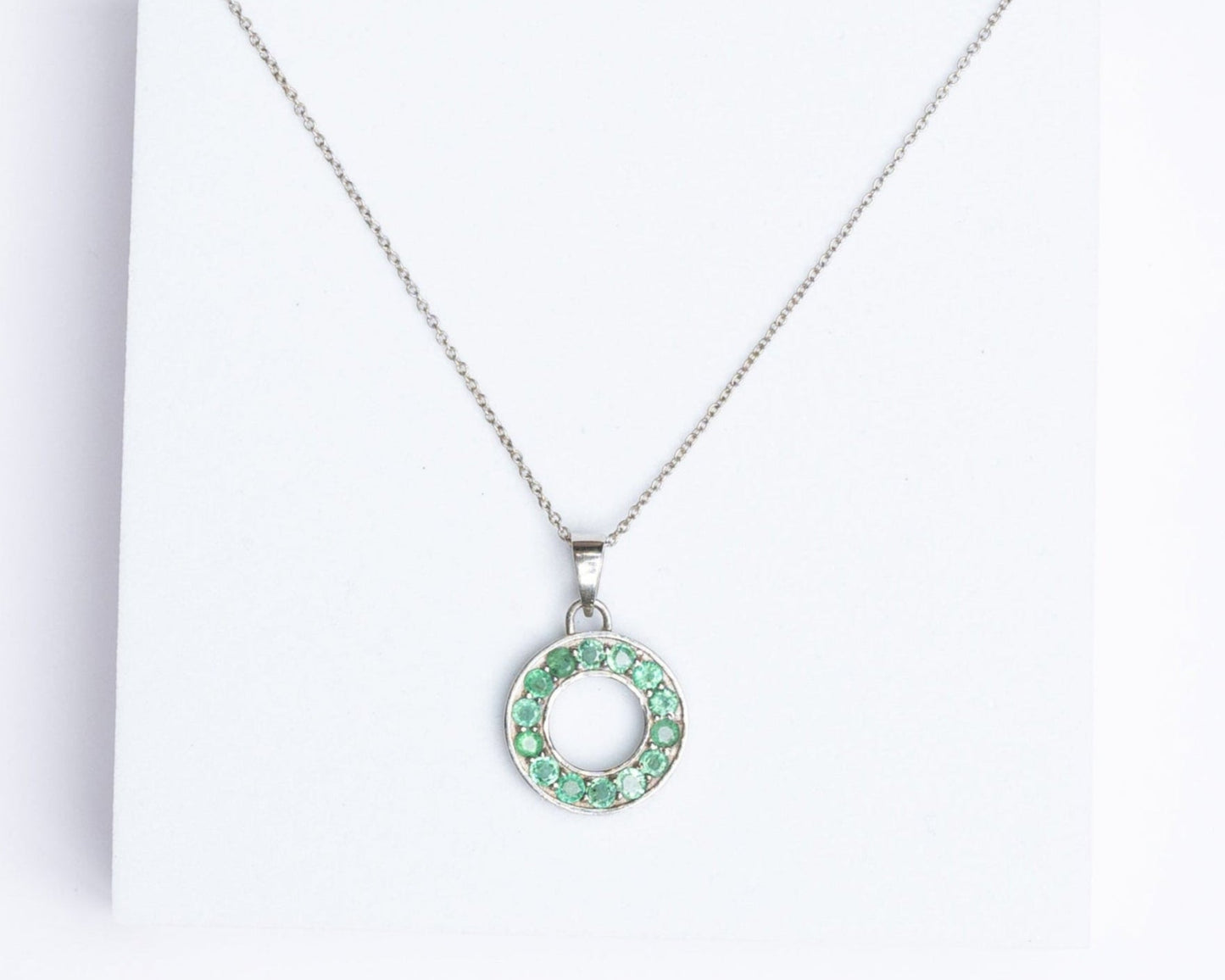 A silver pendant with a round emerald gemstone displayed on a white stand.