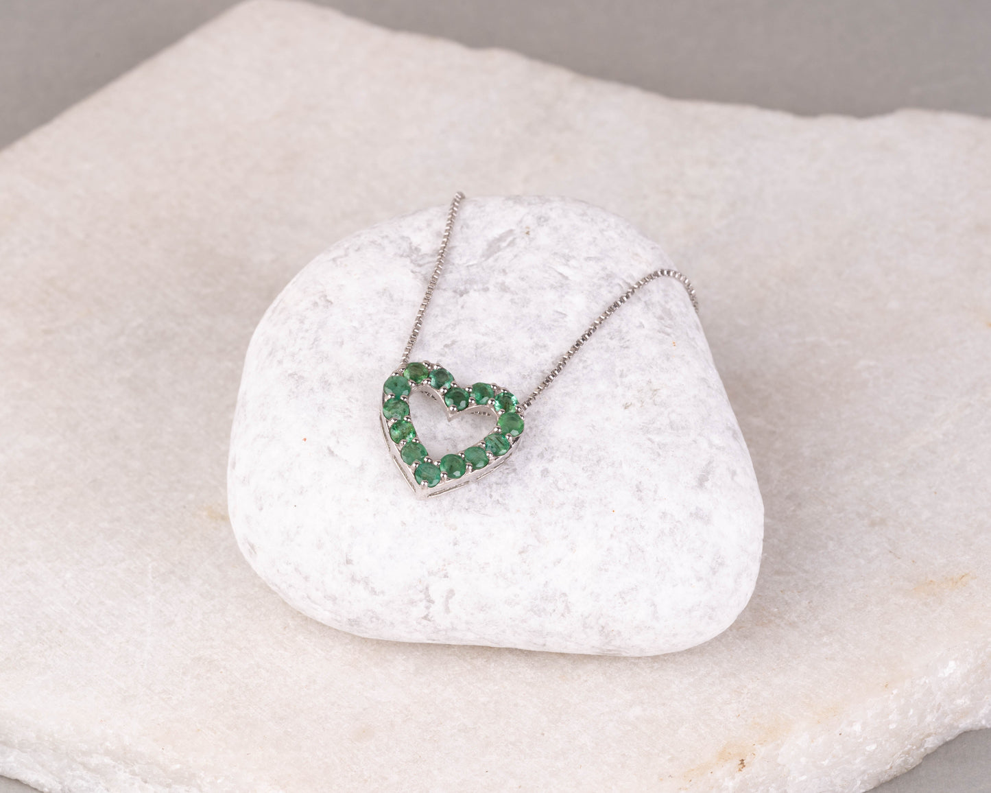 An emerald green gemstone pendant in the shape of a heart, set in a white gold or silver mounting, displayed on a white marble surface.