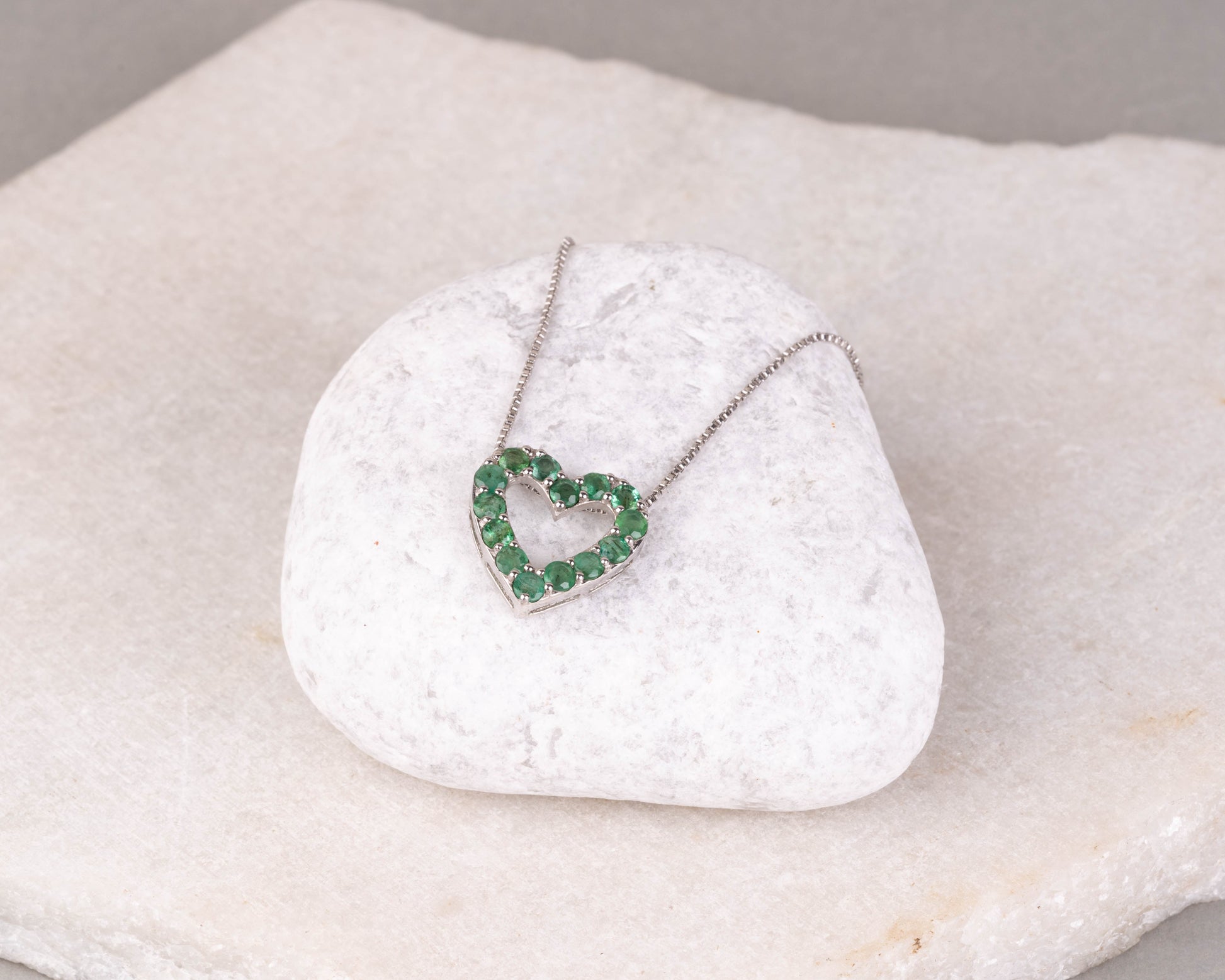 An emerald green gemstone pendant in the shape of a heart, set in a white gold or silver mounting, displayed on a white marble surface.