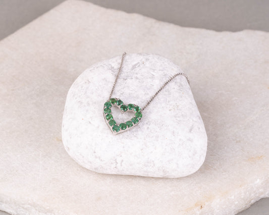An emerald green gemstone pendant in the shape of a heart, set in a white gold or silver mounting, displayed on a white marble surface.